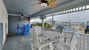 A covered outdoor seating area with white Adirondack chairs and a table, a ceiling fan, and a view of buildings in the background. - Partial beach view home w. shared pool + private elevator. (Tybee Island)