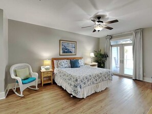 Ein Schlafzimmer mit Bett, Schaukelstuhl, Nachttisch, Deckenventilator, Fenster mit Vorhängen und einem gerahmten Bild an der Wand.
