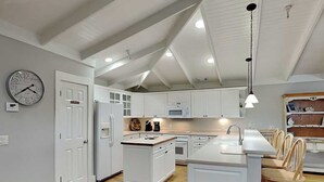 A modern kitchen with white cabinetry, a central island, and a dining area with wooden chairs. - Partial beach view home w. shared pool + private elevator. (Tybee Island)