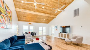 Un salón amplio con techo de madera, un sofá azul, un televisor y un comedor con una mesa y sillas blancas.