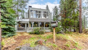 A modern two-story building with large windows, a porch, and a wooden signpost in a forested area. - MELA02 - Spacious Home, Hot Tub, Sauna, Sharc Passes, Dog Friendly (Sunriver)