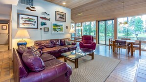 A spacious living room with a leather sofa, a glass coffee table, and a red armchair. There are framed pictures of fish on the wall, and a large window offers a view of the outdoors. - MELA02 - Spacious Home, Hot Tub, Sauna, Sharc Passes, Dog Friendly (Sunriver)