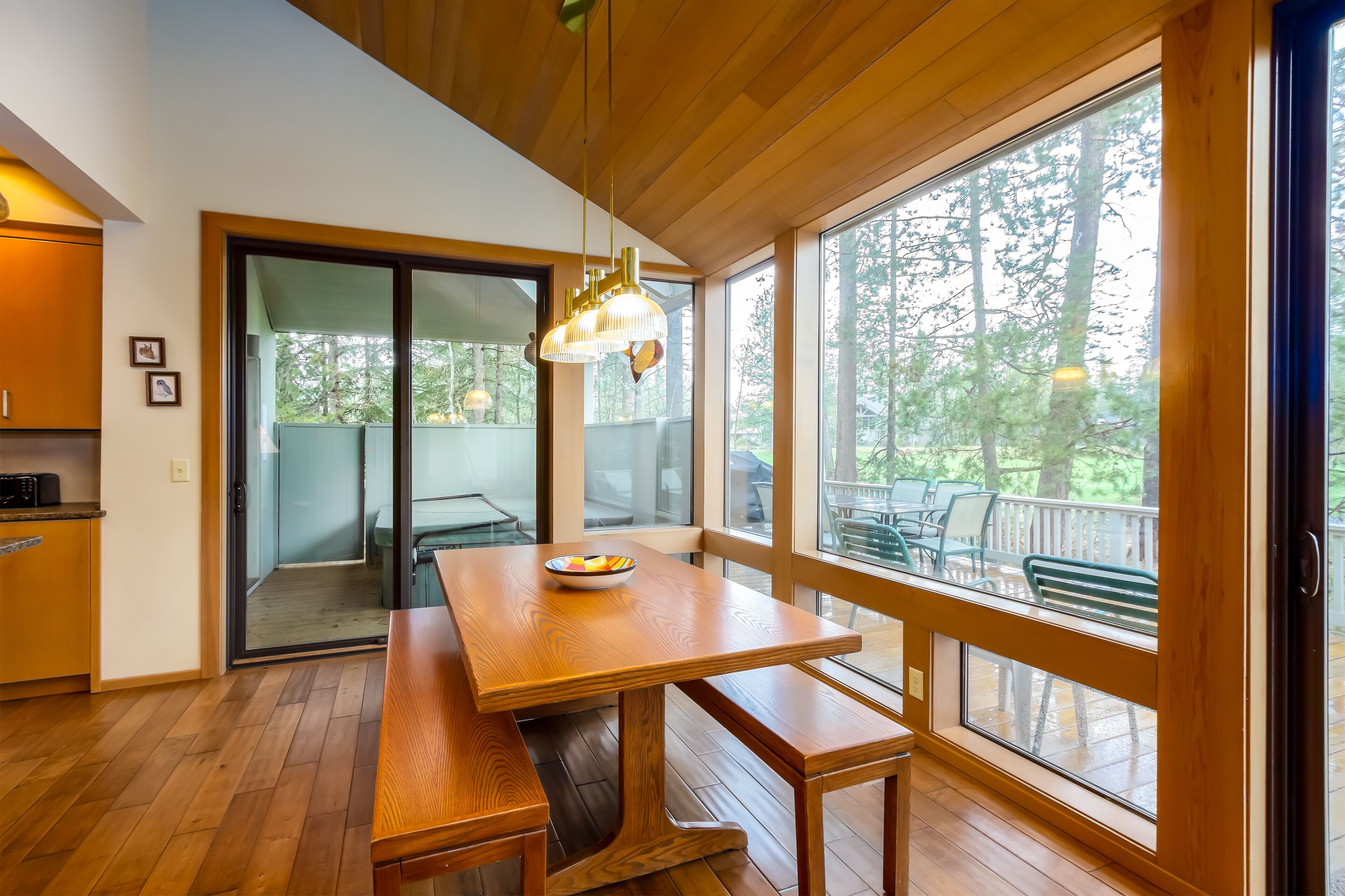 Una mesa de comedor de madera con un cuenco de frutas, ventanas amplias y vistas a árboles.
