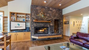 A living room with a stone fireplace, wooden shelves, a leather sofa, and a television displaying 'Bennington Properties'. - MELA02 - Spacious Home, Hot Tub, Sauna, Sharc Passes, Dog Friendly (Sunriver)