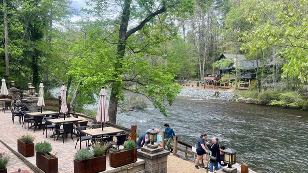 An outdoor patio with tables and chairs, surrounded by greenery and a river.