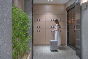 A woman in a white dress with a suitcase in a hallway with wooden lockers and a potted plant.