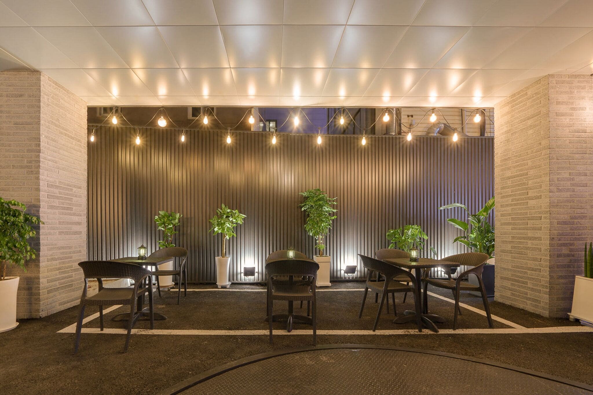 A modern indoor seating area with a brick wall, potted plants, and a ceiling with exposed beams and string lights.
