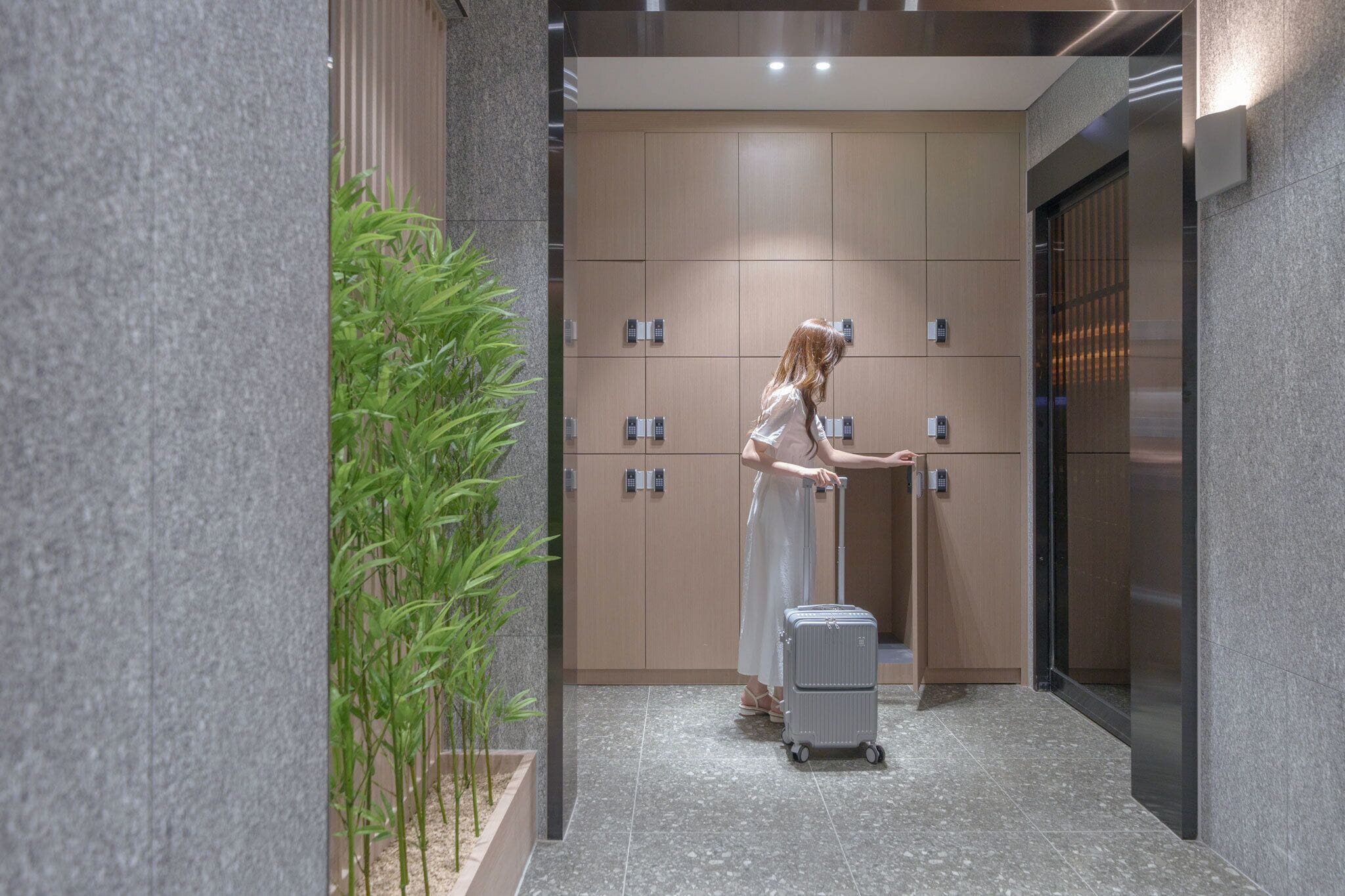Une femme vêtue d’une robe blanche, tenant une valise, se trouve dans un couloir où l’on aperçoit une plante, un ascenseur fixé au mur et une porte dotée d’une poignée.