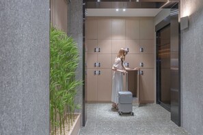 A woman in a white dress with a suitcase in a hallway with a plant, a wall-mounted elevator, and a door with a handle. - Hound Hotel Sungshin Women's University Branch (Seoul)