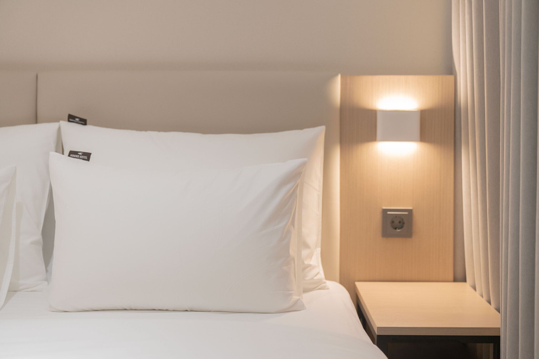 A hotel room with a bed, pillows, a nightstand, and a wall-mounted light fixture.