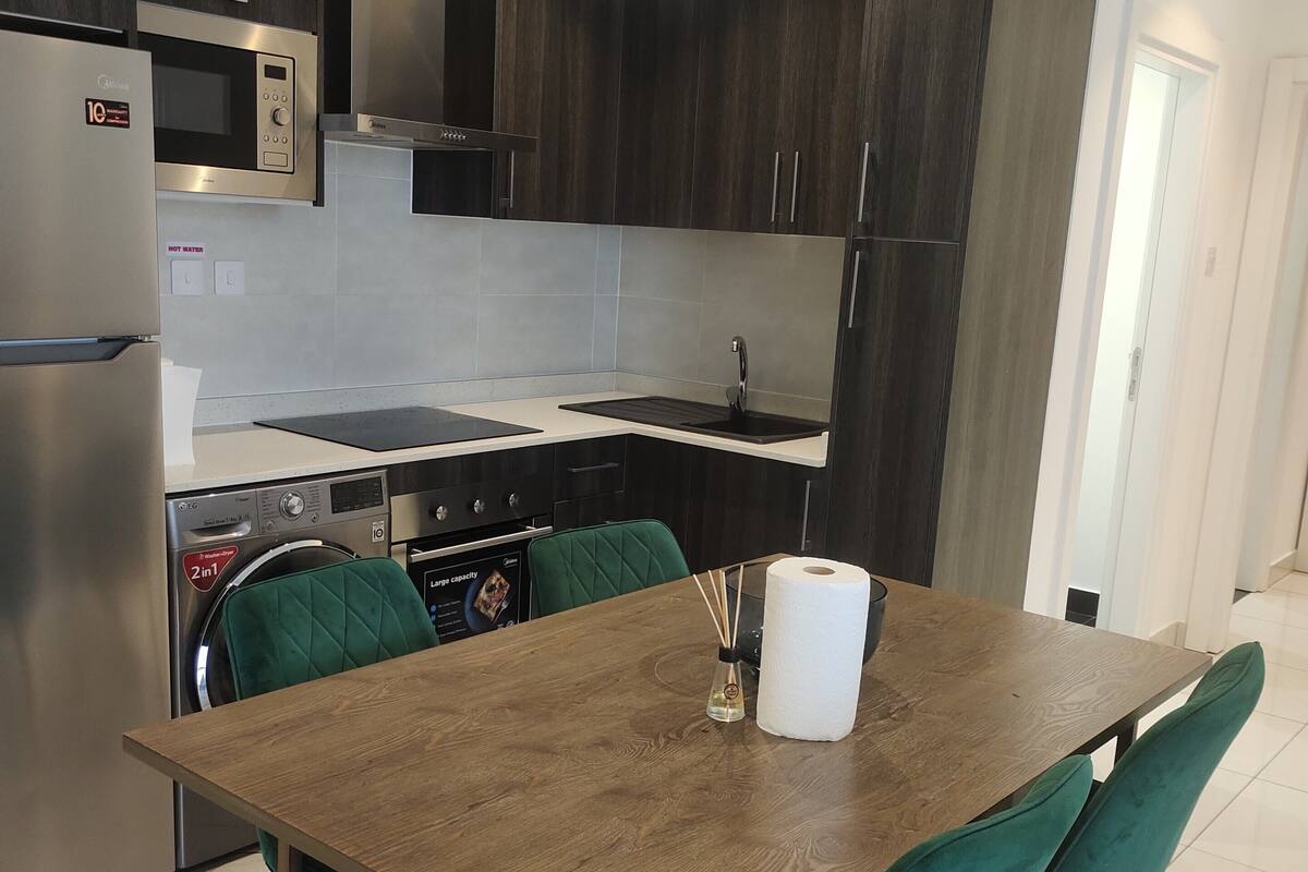 A modern kitchen with dark cabinetry, stainless steel appliances, and a wooden dining table with teal chairs.