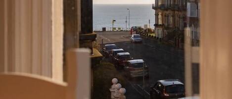 A view through a window of a street with parked cars, a sign for 'Reeds Rains', and buildings with a view of the sea.