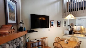 A cozy living room with a fireplace, a dining area with a wooden table and chairs, a TV mounted on the wall, and a kitchen area with a microwave and a hanging light fixture. - Loft in Yosemite National Park (Yosemite West)