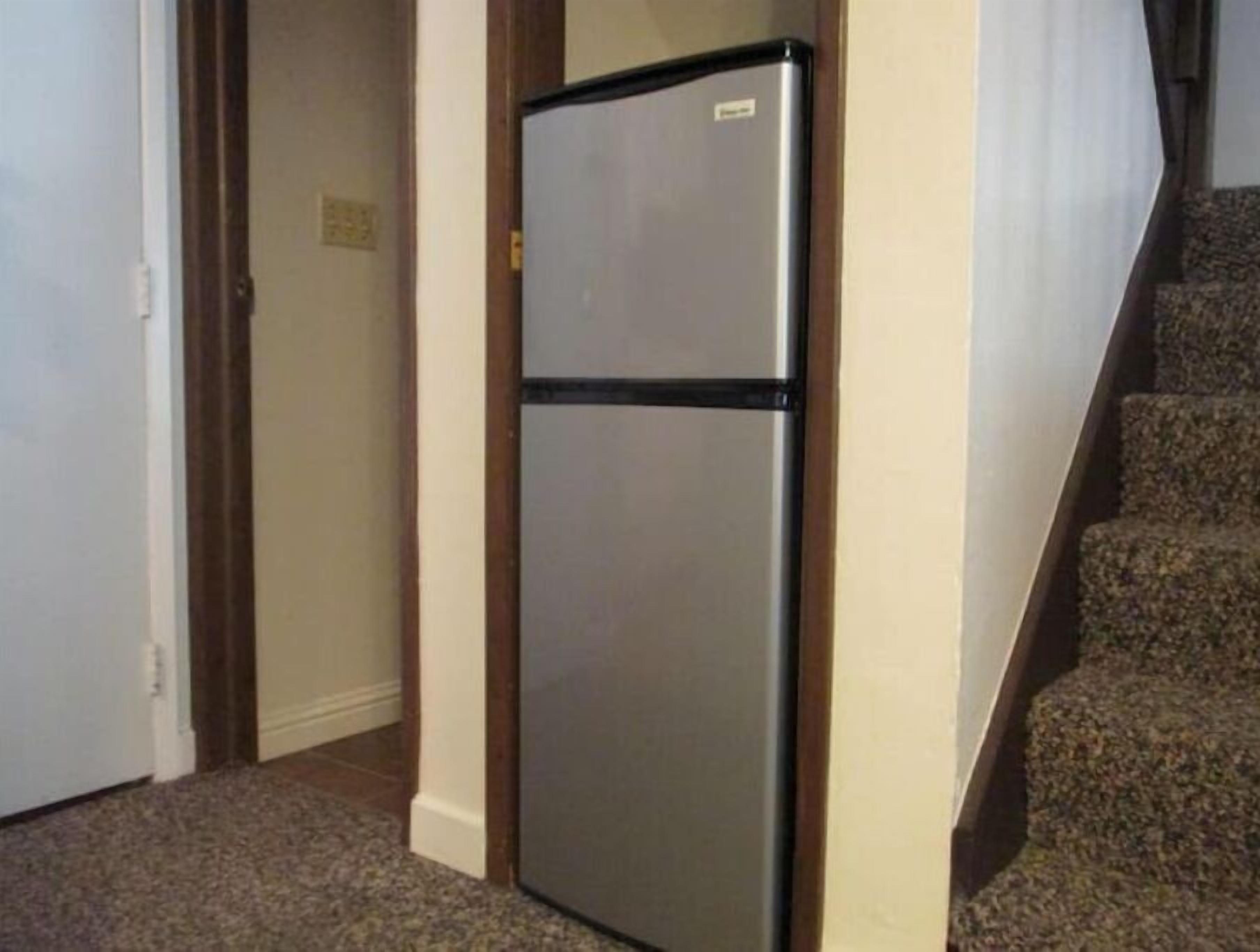 A compact refrigerator in a hallway with carpeted flooring and a wooden door frame.