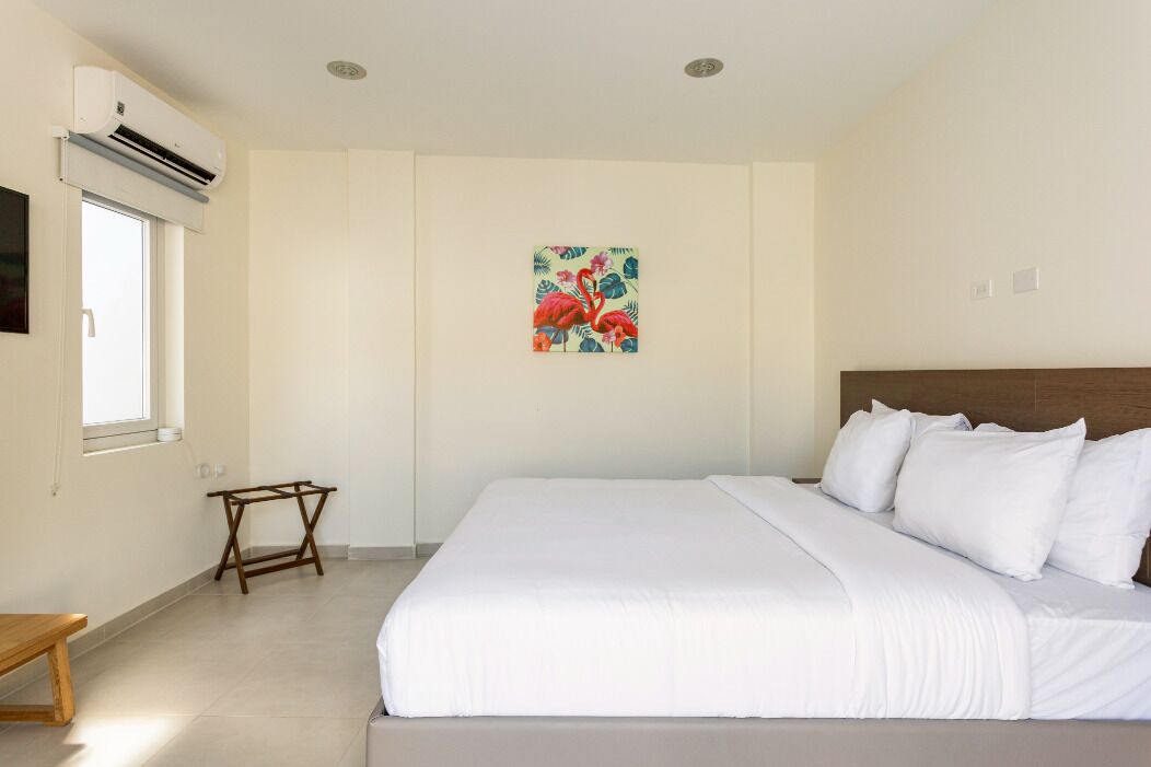 Une chambre dotée d’un grand lit, d’une petite table en bois, d’un appareil de climatisation et d’une seule œuvre d’art accrochée au mur.