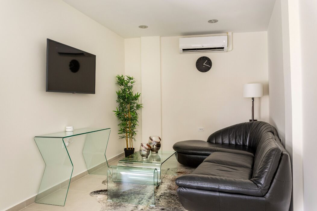 A modern living room with a glass coffee table, a black leather sofa, a potted plant, a wall-mounted clock, and a flat-screen TV.
