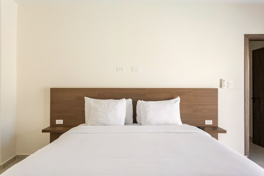 A hotel room with a large bed, two white pillows, a wooden headboard, and a nightstand with a lamp.