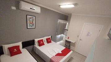 Una habitación de hotel con cama, una mesita, una silla, un equipo de aire acondicionado y una puerta.