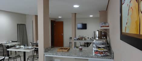 Un comedor moderno con mesas y sillas, una barra de buffet con alimentos y un televisor montado en la pared.