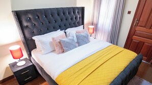 A neatly made bed with a quilted yellow blanket, a tufted headboard, and multiple pillows. There are bedside lamps on either side of the bed. - Silver Harbor Kilimani View Palace Apart (Nairobi)