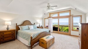 Un dormitorio con una cama grande, mesitas de noche de madera, un aparador, un ventilador de techo y vistas a través de la puerta abierta.