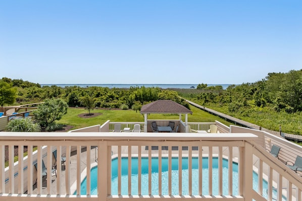 A pool with a gazebo and chairs, surrounded by lush greenery and a view of the ocean.
