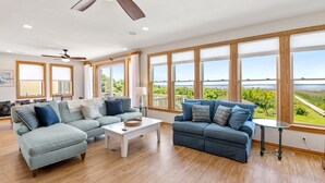Amplia sala de estar con dos sofás, una mesa de centro y un ventilador de techo. Ventanas grandes ofrecen vistas al exterior.