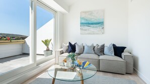 Un soggiorno moderno con un tavolino in vetro, un divano beige con numerosi cuscini, una pianta in vaso e un quadro raffigurante una spiaggia.