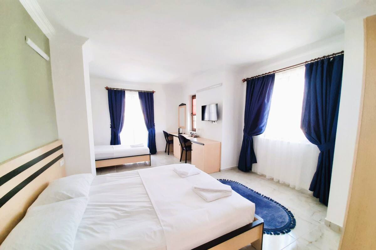 Quarto de hotel com uma cama grande, uma escrivaninha com cadeira, televisão e duas janelas com cortinas.