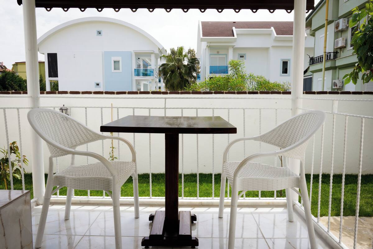 Uma área de estar coberta ao ar livre, com uma mesa e duas cadeiras, cerca branca e edifícios ao fundo.