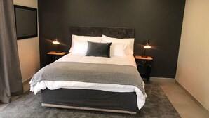 A modern bedroom with a large bed, two bedside tables with lamps, a dark headboard, and a grey carpet. - Exquisite one bedroomed apartment - 2043 (Bulawayo)