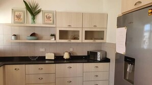 Una cocina moderna con armarios de madera clara, una encimera negra, un refrigerador de acero inoxidable, un microondas y un dispensador de agua.