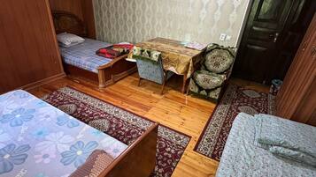 A room with two beds, a small table, and a chair. The room has patterned wallpaper and wooden flooring.