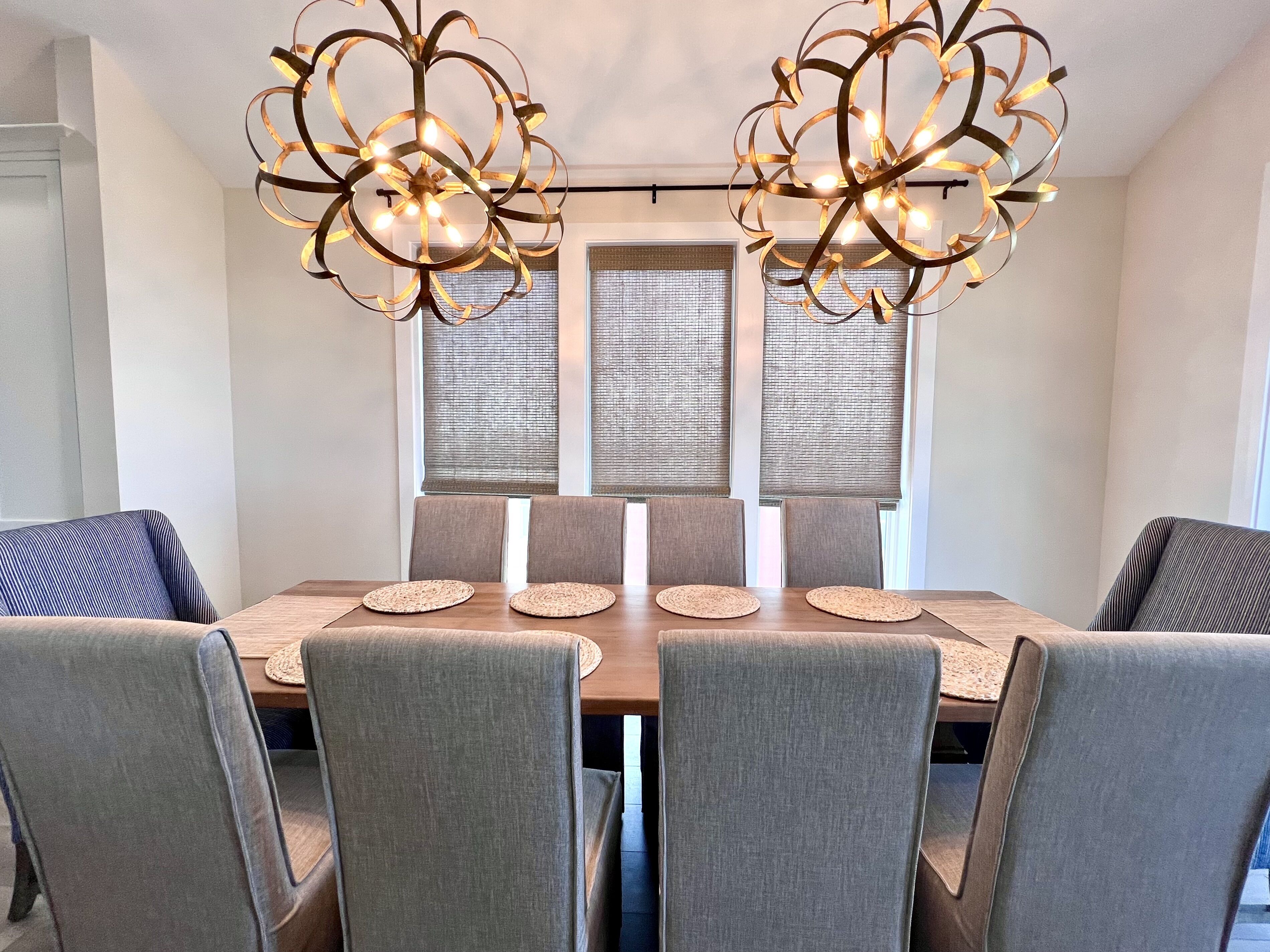 A dining area with a wooden table, six grey upholstered chairs, and two modern chandeliers.