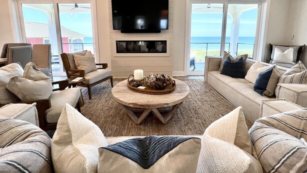 A spacious living room with a large flat-screen TV mounted on the wall, a cozy seating area with multiple sofas and chairs, a wooden coffee table, and large windows offering a view of the ocean.