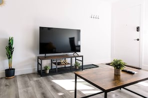 A modern living room with a flat-screen TV on a wooden stand, a coffee table, and two potted plants. - The Cozy Crib / Spacious & Workspace w/ Monitor (Salt Lake City)