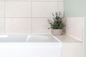 Um banheiro com uma banheira branca, uma planta de lavanda em vaso no balcão e paredes com azulejos bege.