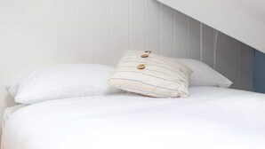 Een slaapkamer met een schuine kap, wit beddengoed en gestreepte kussens met knopen.