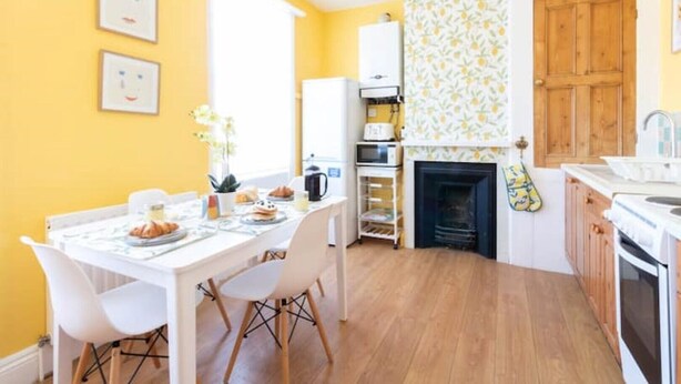 Een heldergele kamer met een ontbijttafel, een wit kastje en een open haard met een decoratieve bovenkant.
