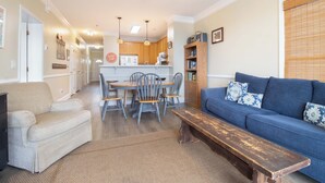 Un salon avec un canapé, un banc, une table à manger avec des chaises, une cuisine à l’arrière-plan et un ventilateur de plafond.