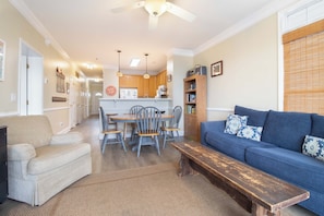 A living room with a sofa, a bench, a dining table with chairs, a kitchen in the background, and a ceiling fan.