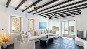 A modern living room with a large beige sectional sofa, a black coffee table, and a ceiling fan. There are two windows with stone frames, a painting on the wall, and a TV on a white stand. - Luxury Villa Agavi Ideal for Events (Mykonos)