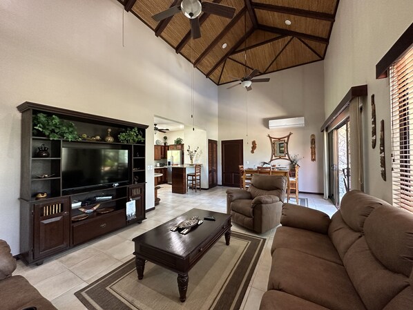 A spacious living room with a large flat-screen TV, dark wood furniture, a ceiling fan, and a kitchen area in the background. - The perfect home away from home (Playas del Coco)
