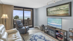 Un salon avec un canapé, un fauteuil, un téléviseur et un tableau représentant une scène de plage.