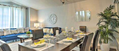 Comedor con una mesa de madera dispuesta para seis comensales, una araña, un reloj y un espejo grande en la pared.