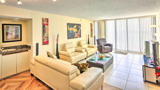 A modern living room with a sofa, armchairs, a coffee table, and a TV stand. There is a kitchen area with a sink and cabinets. The room has a large window with blinds and a ceiling light fixture. - Bayfront Miami Condo w/ Resort Perks & Ocean Views (Miami)