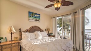 A bedroom with a large bed, a ceiling fan, a nightstand with a lamp, a painting of a beach scene, and a view of the ocean through a sliding glass door. - Best Views in St Pete for Dolphin Viewing (St. Petersburg)