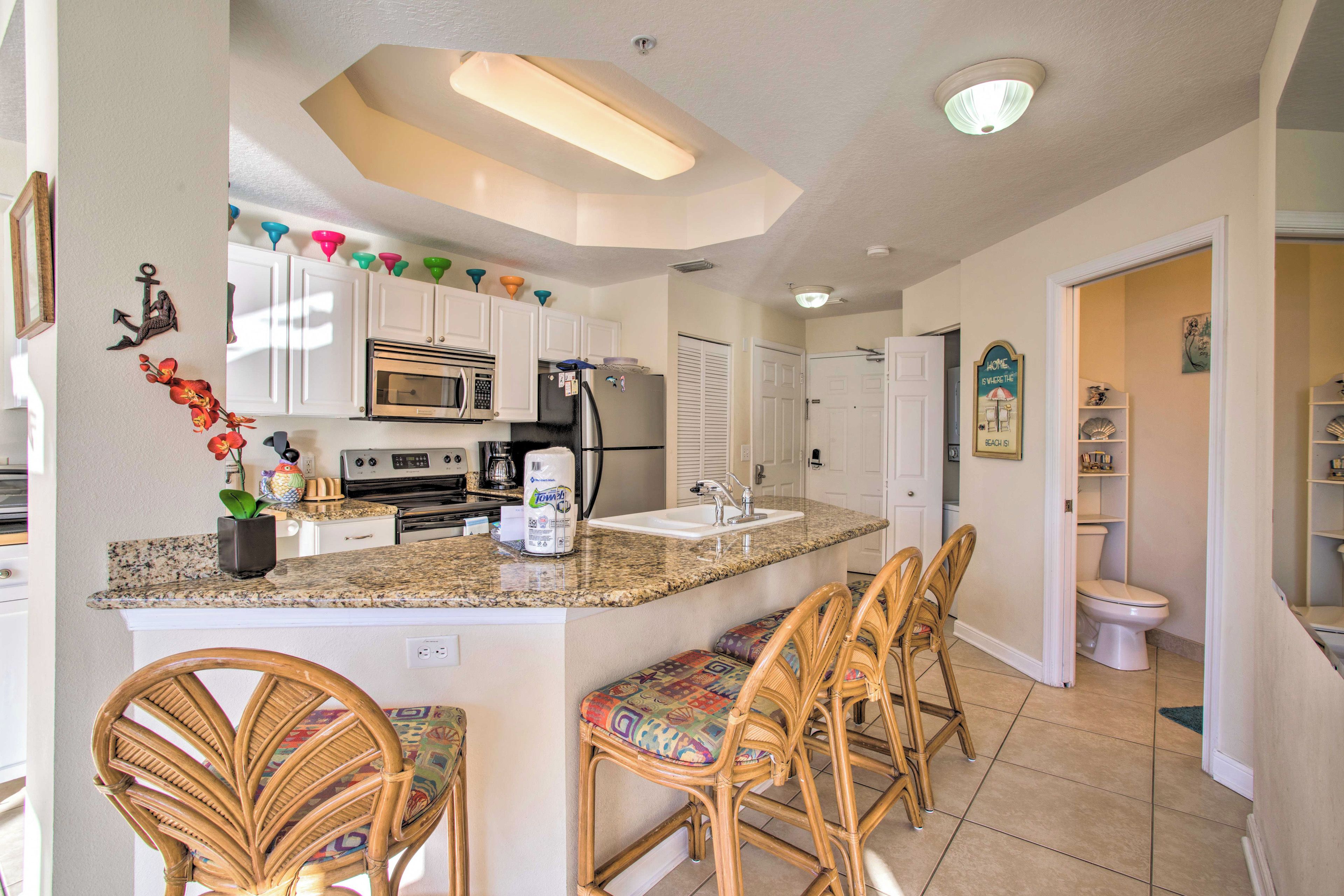 A kitchen with white cabinets, a granite countertop, a microwave, a refrigerator, and a sink. There are wicker chairs with colorful cushions.