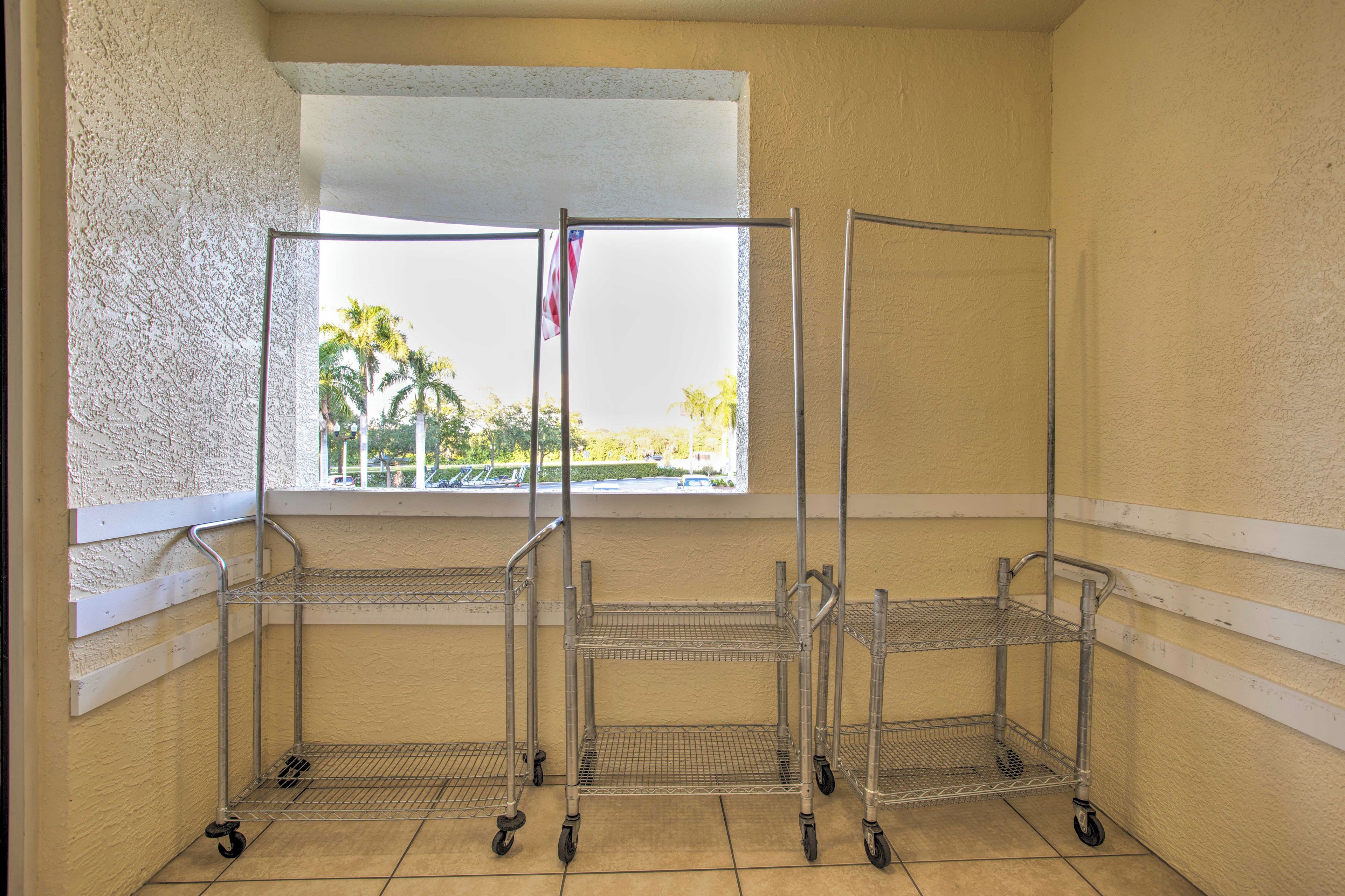 A corner of a room with three metal wire shelving units against a wall, a window with a view of palm trees, and a white baseboard.