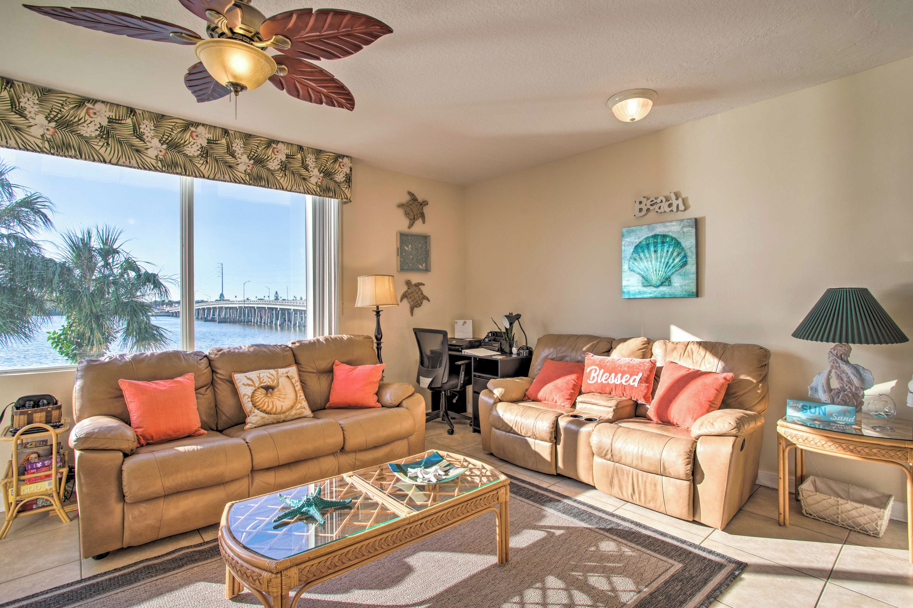 A living room with two leather sofas, a glass coffee table, and a ceiling fan. There is a desk with a computer and a lamp. The room has large windows with a view of palm trees and a pier.